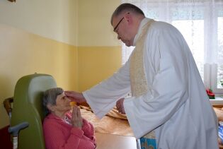 XXIX Światowy Dzień Chorego 11.02.2021 pokój w domu, a w nim kobieta siedząca w fotelu rehabilitacyjnym oraz kapłan udzielający jej sakramentu namaszczenia chorych.