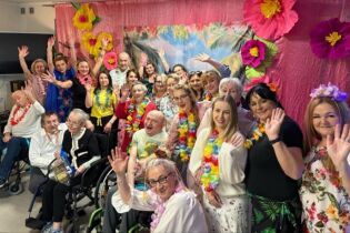 Aloha!!! duża grupa osób machających rękoma. Większość z nich na szyi ma girlandy z kwiatów w stylu hawajskim