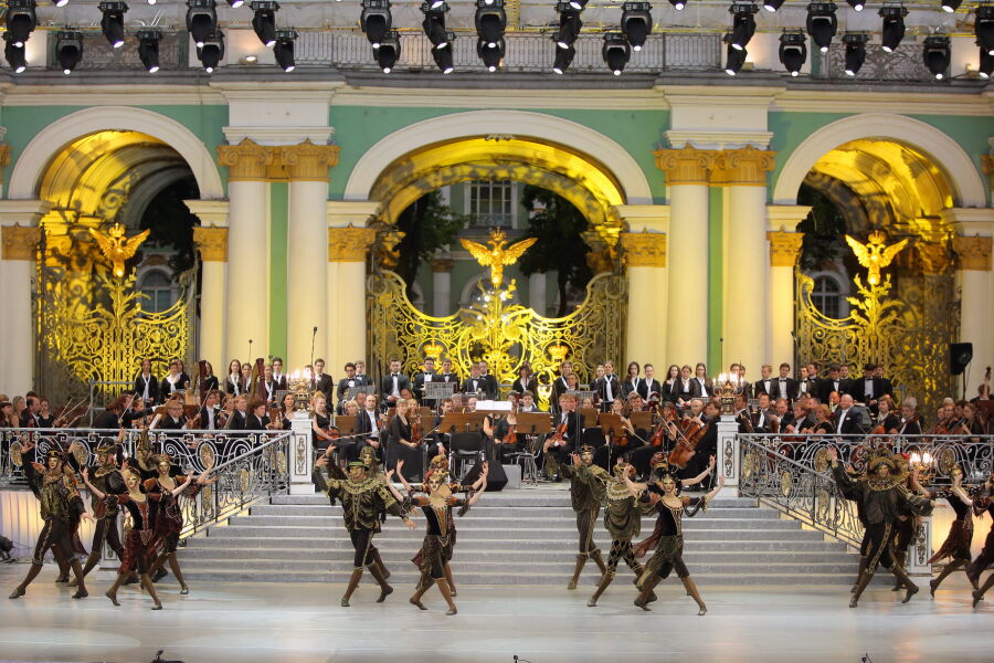 Na tle zielonej zabytkowej fasady Pałacu na Placu Pałacowym siedzi orkiestra symfoniczna. Na pierwszym planie pary tancerzy w strojach Arlekinów i maskach na twarzach