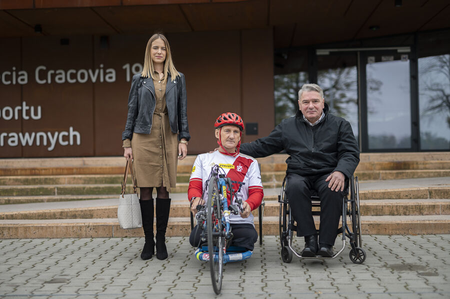 trzy osoby stojące w rzędzie na tle budynku Centrum sportu niepełnosprawnych. Kobieta z lewej strony, kolarz na rowerze ręcznym w środku, z lewej strony mężczyzna na wózku inwalidzkim.