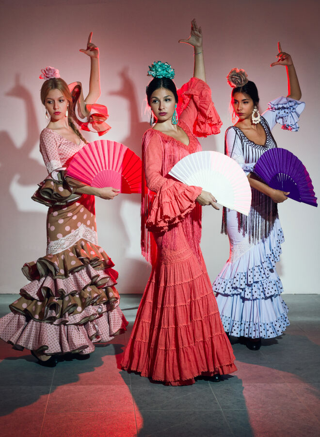 Kultura Hiszpanii - taniec Flamenc. Trzy tancerki Flamenco w charakterystycznych sukienkach z falbanami u dołu spódnicy trzymają wachlarze. Jedną rękę mają uniesioną do góry jak w tańcu 