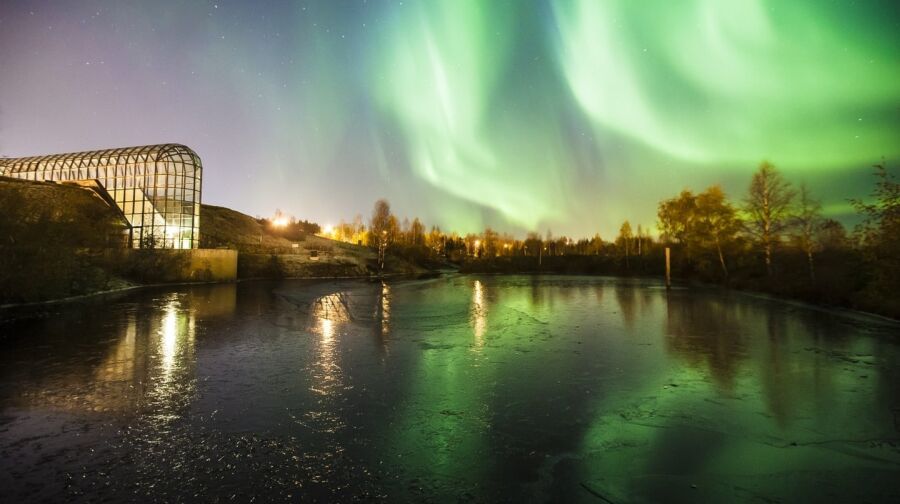 Zorza polarna w Finlandii. Zbiornik wodny i oświetlona konstrukcja ze szkła i stali. Wokół zbiornika wodnego drzewa. Na ciemniejszym  niebie jasnozielone świetliste pasy zorzy polarnej.  