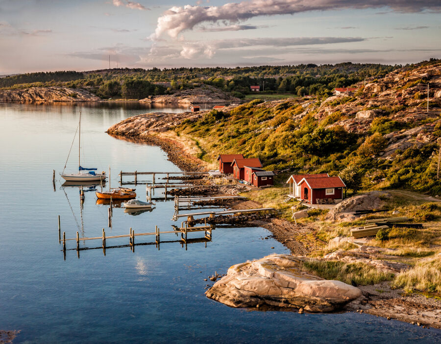 Zachodnie wybrzeże Szwecji. Skaliste wybrzeże i zatoka po lewej kilka małych kolorowych domków oraz drewniane przystanie z kilkoma niewielkimi łodziami 