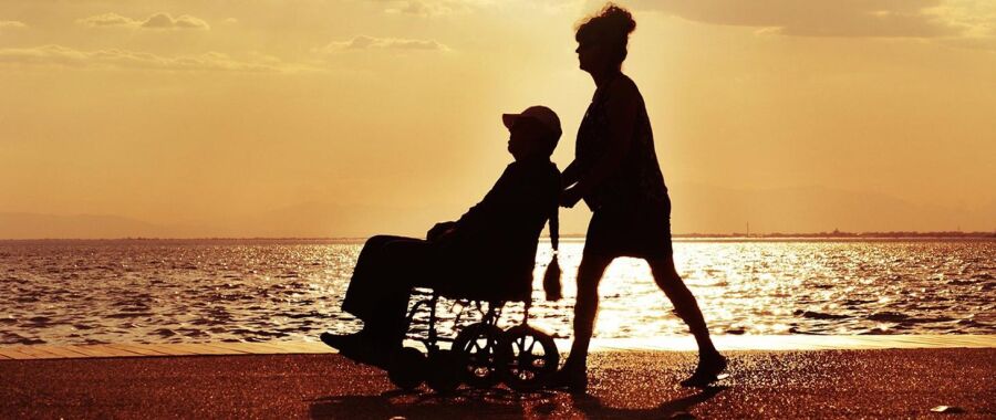 Zdjęcie przedstawia osobę na wózku inwalidzkim z drugą osobą pchającą ten wózek na plaży i tle zachodzącego słońca