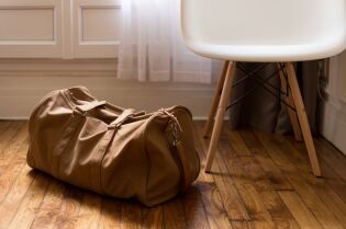 Grafika przedstawia torbę podróżną, stojącą na podłodze obok krzesła.