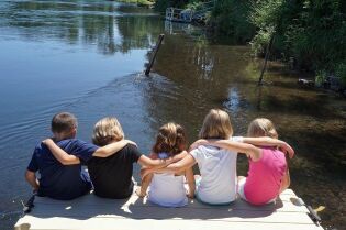 Grafika przedstawia pięcioro dzieci siedzących na pomoście nad wodą. Dzieci widoczne są od tyłu.. 