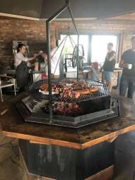 W pomieszczeniu restauracyjnym z oknem na ośmiokątnym grillu nad paleniskiem leżą kiełbasy i szaszłyki. Nad grillem unosi się dym. Na dalszym planie kelnerka obsługuje klienta oraz stoją inne osoby.