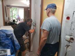 Ściany z widoczną instalacją elektryczną, pochylony mężczyzna w ubraniu roboczym instaluje kontakt elektryczny, przy nim drugi Pan odwrócony tyłem, widoczne meble zabezpieczone folią malarską.