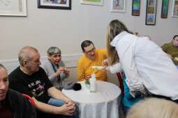 Grupa kobiet i mężczyzn siedzących w pomieszczeniu przy nakrytym stole. Na pierwszym planie kobieta i mężczyzna częstujący się jajkami. 