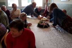 Grupa kobiet i mężczyzn siedzących w pomieszczeniu przy nakrytym stole, na którym znajduje się świątecznie udekorowany wianek. W tle mężczyźni częstują się jajkami. 