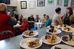 Grupa kobiet i mężczyzn w pomieszczeniu. Na pierwszym planie blat z talerzami, na których wyłożone są kawałki ciast. 