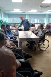 Grupa kobiet i mężczyzn siedzących w pomieszczeniu przy stołach. W tle mężczyźni podają sobie ręce.