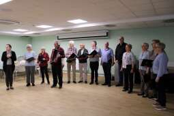 Grupa kobiet i mężczyzn z nutami w dłoniach w pomieszczeniu. Na pierwszym planie przemawiająca kobieta i mężczyzna z akordeonem.