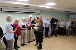 Grupa stojących kobiet i mężczyzn w pomieszczeniu. Dwie kobiety wręczają podarunki muzykom.