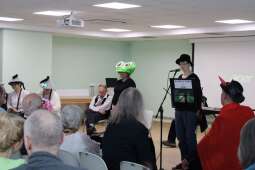 Grupa kobiet i mężczyzn ujętych tyłem. Pośrodku kilka osób występujących w kostiumach teatralnych.