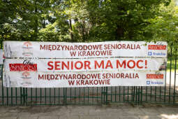 Zdjęcie zawiera baner z XII Międzynarodowych Senioraliów w Krakowie zawieszony na płocie.