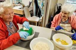 Na zdjęciu uśmiechnięte Mieszkanki, jedna trze ser żółty, druga kroi paprykę. 