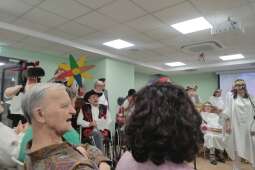 Na zdjęciu na pierwszym planie mieszkaniec włącza się w śpiew, w tle grupa przedstawiająca program Jasełek w trakcie śpiewania kolęd. 