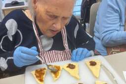 Na zdjęciu Mieszkanka smaruje przygotowane fragmenty ciasta drożdżowego dżemem. 