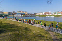 Biegacze z numerami startowymi biegną bulwarami wiślanymi w słoneczny dzień. Trasa prowadzi wzdłuż rzeki Wisły, w tle widoczna woda, zieleń oraz zabudowania. Uczestnicy poruszają się zwartą grupą.
