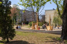 Widać dzieci i dorosłych, korzystających z nowo otwartej przestrzeni ogrodu. W tle znajduje się plac z zielenią, nowymi nasadzeniami oraz elementami małej architektury i atrakcjami.