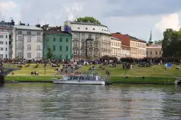Ludzie odpoczywają na trawie nad rzeką, w tle budynki i łódź.