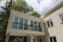 Balkon na elewacji budynku z dużymi oknami i otaczającą zielenią.