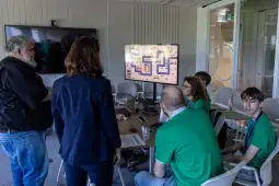 Grupa osób pracuje nad projektem przy stole, z ekranem pokazującym grę komputerową.