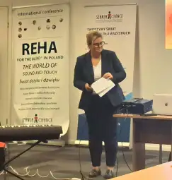 Kobieta w garniturze prezentuje na konferencji, obok instrument muzyczny i materiały informacyjne.