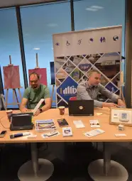 Dwa mężczyźni przy stole z laptopami, materiałami promocyjnymi i plakatem w tle.