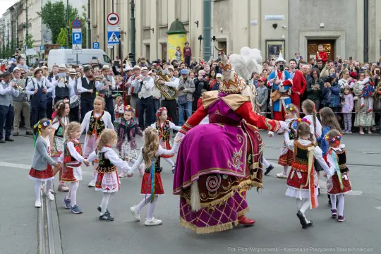 Grupa dzieci w tradycyjnych strojach tańczy w korowodzie z dorosłymi w tle.