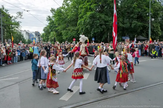 Uczestnicy parady w tradycyjnych strojach ludowych tańczą na ulicy, otoczeni tłumem widzów.
