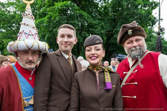 Cztery osoby w strojach historycznych, uśmiechające się na tle zieleni.