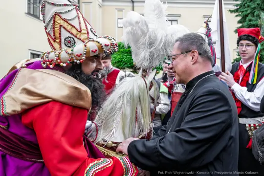 Mężczyzna w tradycyjnym stroju rozmawia z innym mężczyzną w garniturze. W tle widoczni uczestnicy wydarzenia.