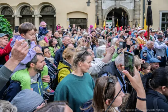 Tłum ludzi z telefonami, niektórzy trzymają dzieci, w tle flagi i postacie w strojach historycznych.