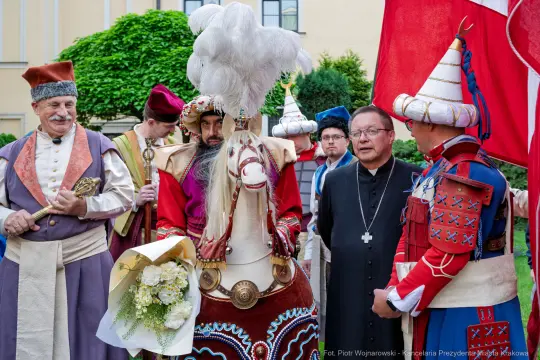 Grupa osób w tradycyjnych strojach, w tym jeździec na koniu, z kwiatami w ręku.
