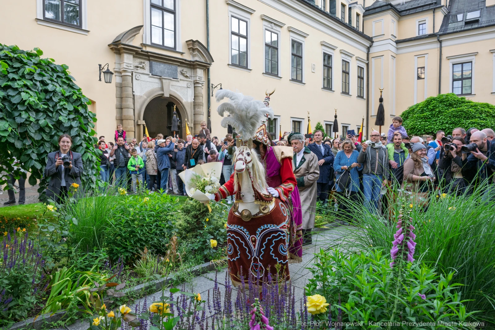 Kobieta w tradycyjnym stroju z kwiatami, prowadząca procesję wśród tłumu ludzi w ogrodzie.  Autor: Piotr Wojnarowski
