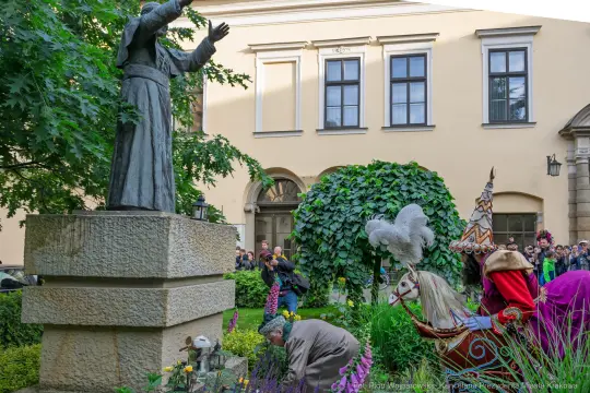 Statua świętego, ludzie w strojach ludowych, dekoracje kwiatowe w ogrodzie.