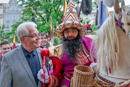Mężczyzna w kolorowym stroju z koszem i berłem, obok niego inny mężczyzna w garniturze.