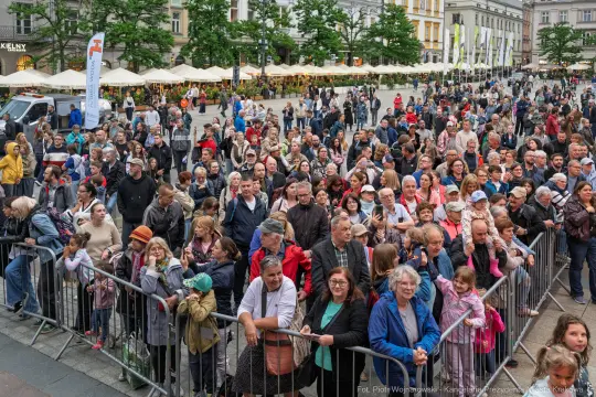 Tłum ludzi zgromadzony na placu, z widocznymi straganami i drzewami w tle.