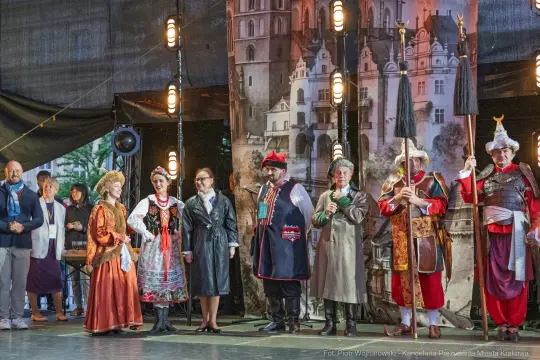 Grupa osób w tradycyjnych strojach ludowych na scenie, w tle architektura.