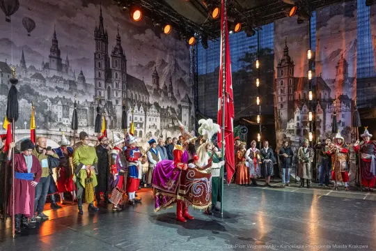 Uroczystość z udziałem postaci w strojach historycznych na tle malowniczej scenografii.