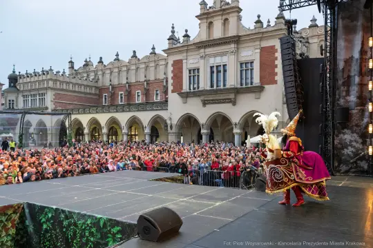 Tłum ludzi na placu, występ artystyczny z kolorowymi strojami i dekoracjami.