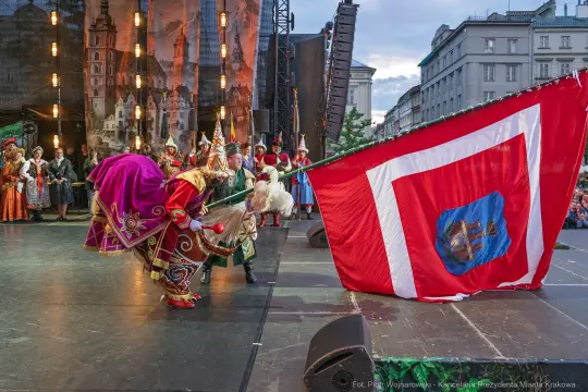 Tradycyjny występ z tańcem i flagą w tle, na scenie w Krakowie.