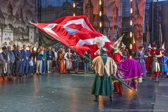 Tradycyjny występ z flagą, uczestnicy w strojach ludowych, tło z architekturą.