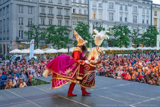 Kostiumowany artysta w kolorowej szacie tańczy na scenie przed tłumem ludzi.