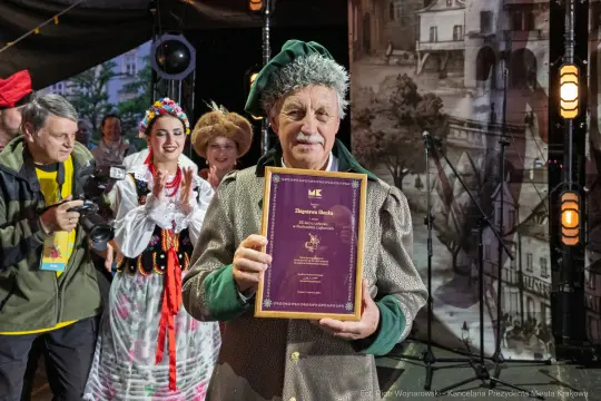Mężczyzna w stroju ludowym trzyma dyplom, obok niego tańczące kobiety w tradycyjnych strojach.