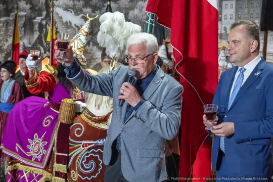 Mężczyzna w szarym garniturze wznosi toast, obok niego stoi drugi mężczyzna. W tle postacie w strojach ludowych.