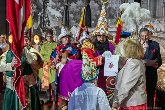 Uczestnicy wydarzenia w kolorowych strojach, wręczający nagrody i trzymający flagi.