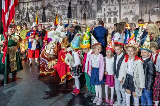 Grupa ludzi w tradycyjnych strojach, dzieci w kolorowych czapkach, podczas uroczystości.